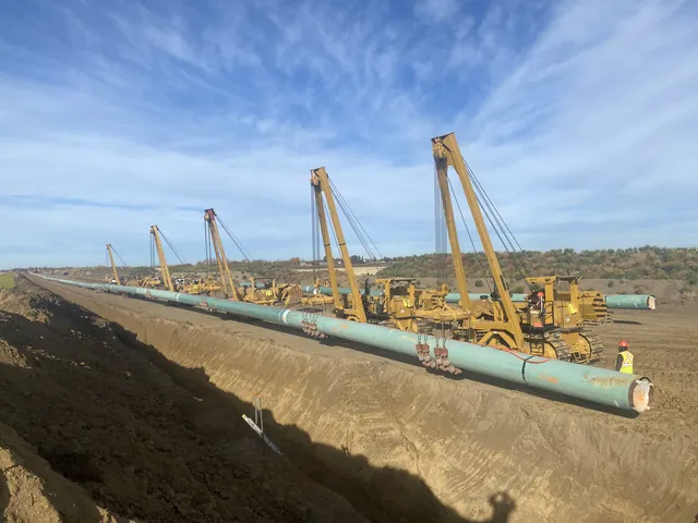A line of Midwest pipelayers supporting a long section of green-coated pipe alongside an open trench