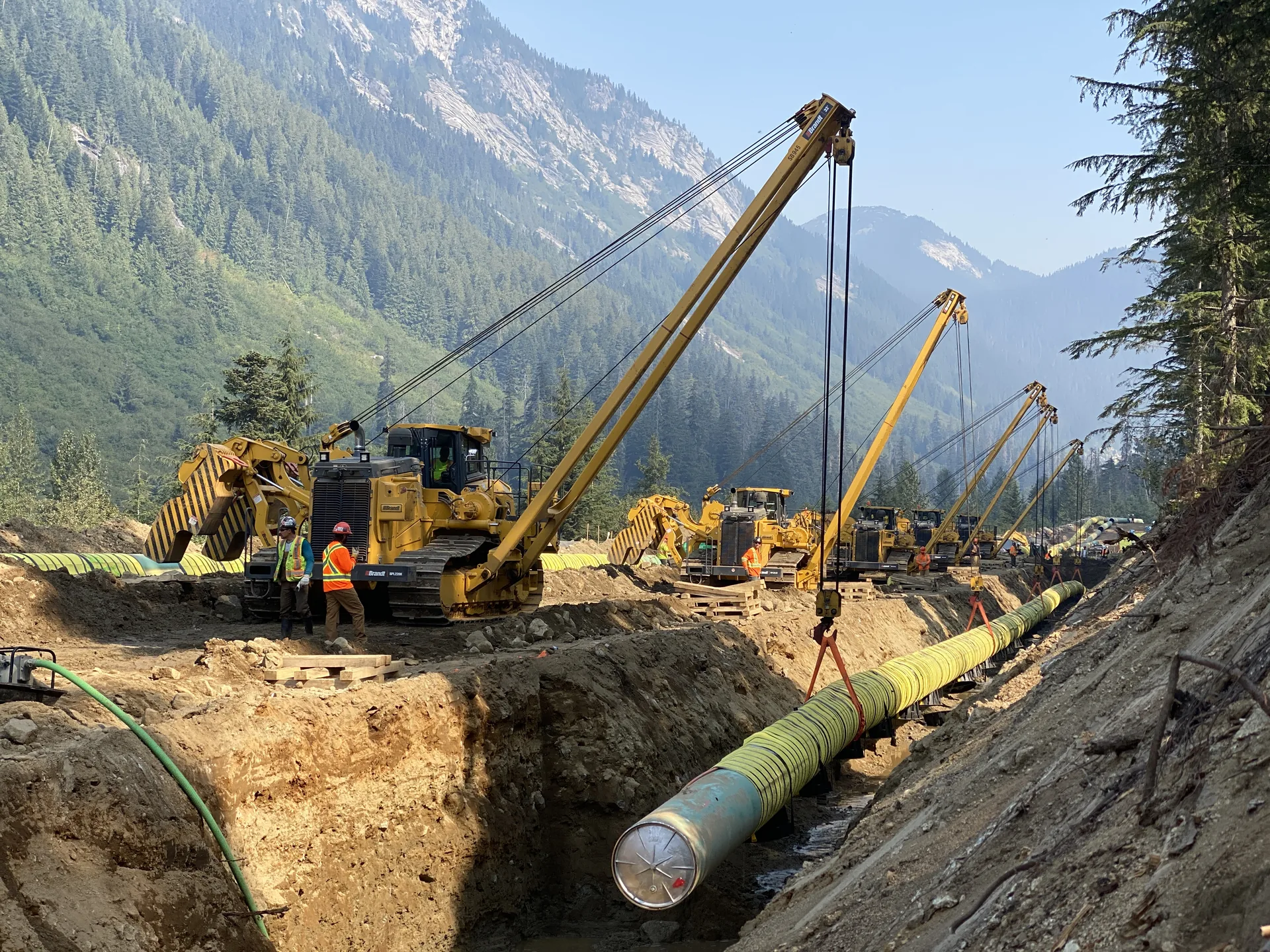 Midwest crews and a line of pipelayers lowering pipe through a mountain construction corridor